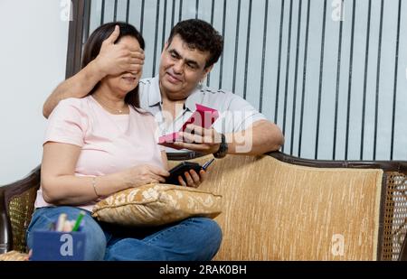 Indian husband covering his wife eyes, making birthday surprise, giving her gift at home. Millennial diverse couple celebrating b-day together, enjoyi Stock Photo