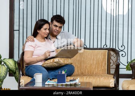 Portrait of young Indian happy cheerful family couple resting on sofa in living room searching internet typing online on laptop computer and speaking, Stock Photo