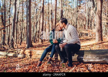 Happy loving couple drinking tea from thermos relaxing in autumn forest. Man and woman sitting on trunk having coffee from vacuum flask. People kissin Stock Photo