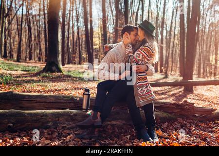 Portrait of young loving couple drinking tea from thermos relaxing in fall forest. Man and woman sitting on trunk hugging enjoying autumn landscape Stock Photo