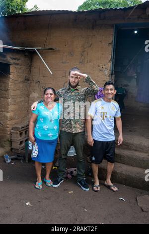 Ahuachapán, El Salvador - November 04 2022: Salvadoran Woman Hugging a Tall White Blond Man in Military Clothes next to a Dark-Haired Man Outside her Stock Photo