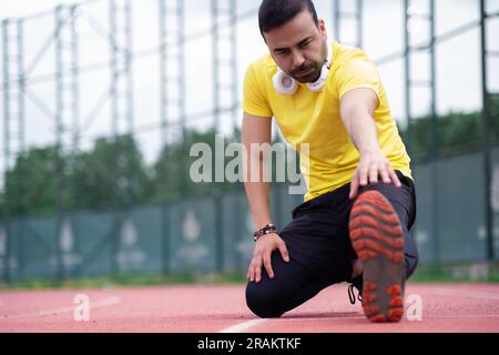 Man with headphones in activewear stretching leg sitting and bending forward on red running track Stock Photo