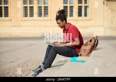 Male Student Working On Laptop Sitting On StepsOutside University Building In Oxford UK Stock Photo