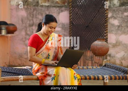 Happy Indian village woman in saree using the laptop sitting outside the house doing thumbs up. Rural Indian woman sitting on cot. technology concept. Stock Photo