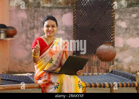 Happy Indian village woman in saree using the laptop sitting outside the house doing thumbs up. Rural Indian woman sitting on cot. technology concept. Stock Photo