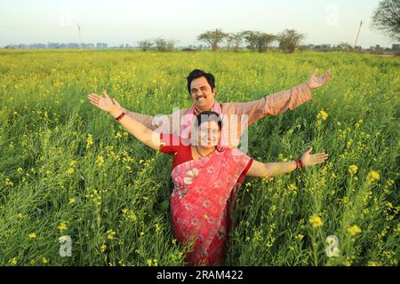 Happy Indian farmer couple standing in mustard field enjoying the agricultural profits and happy to see the flourished agricultural crops benefits. Stock Photo