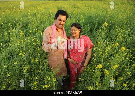 Happy Indian farmer couple standing in mustard field enjoying the agricultural profits. Money in hand. Happy to get benefitted by mustard crops. Stock Photo