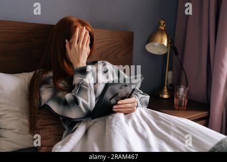 Side view of grief-stricken young woman lying on bed covering face with hand and crying while holding picture frame, touching photograph with love. Stock Photo