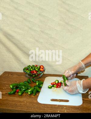 Wearing plastic gloves, the kitchen worker holds the jalapeno pepper and carefully slices it. Stock Photo