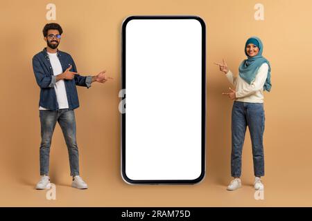 Nice Offer. Smiling Muslim Couple Pointing At Blank Smartphone With White Screen Stock Photo