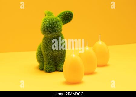 Green Easter Bunny with three yellow candles of eggs shape on matching monochrome orange background. Minimal Happy Easter holiday concept. Creative Stock Photo