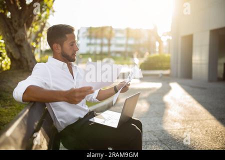 Man business analyst, accountant sitting on bench with laptop, holding papers, checking correspondence outdoors Stock Photo