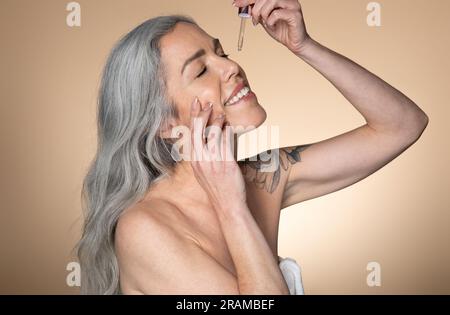 Beautiful smiling senior woman applying facial serum, using anti-aging treatment product, beige studio background Stock Photo