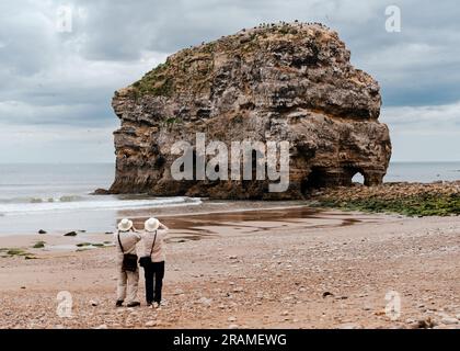 Two retired people using binoculars to view the bird colony on the top of Marsden Rock on Marsden Beach in South Shields. Stock Photo