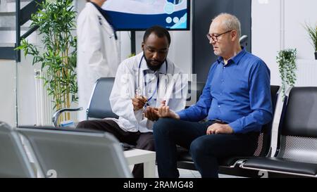 Specialist doing medical test with diabetic man in hospital waiting room, measuring insulin level and glucose in blood sample using glucometer instrument. Elderly man having checkup visit appointment Stock Photo