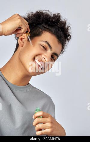 Young guy with black curly hair moisturizing his skin with a facial oil in an anti-aging skincare routine. Handsome swarthy man pampering his skin to Stock Photo