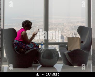 A woman looks at her telephone at a window in the Burj Khalifa building overlooking Dubai, UAE Stock Photo