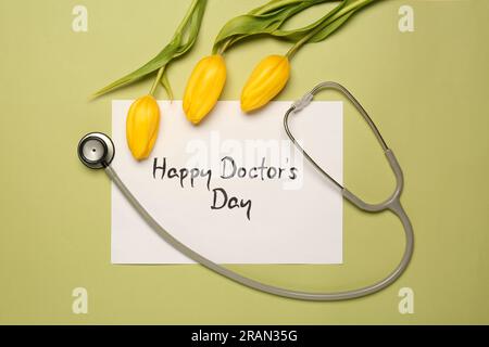 Card with phrase Happy Doctor's Day, stethoscope and yellow tulips on light green background, flat lay Stock Photo