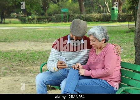 Old age couple sitting on the bench in a lush green serene environment having a good time together. They are happily enjoying their time. Stock Photo