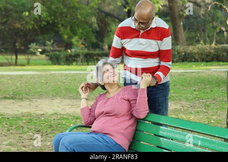 Old age couple sitting on the bench in a lush green serene environment having a good time together. They are happily enjoying their time. Stock Photo