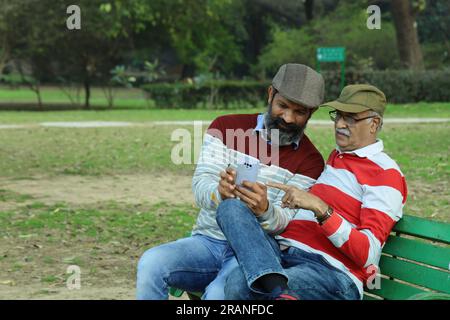Old father and mid aged son sitting on park bench holding mobile phone in hand. Video call and taking selfie. They are in lush greens and serenity. Stock Photo