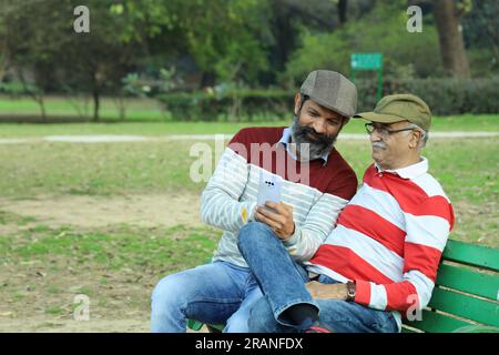 Old father and mid aged son sitting on park bench holding mobile phone in hand. Video call and taking selfie. They are in lush greens and serenity. Stock Photo