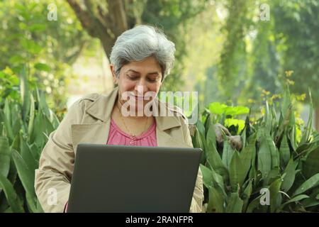 Old confident woman sitting in lush green atmosphere holding laptop. working online, making best use of technology and internet. Self-dependent woman. Stock Photo