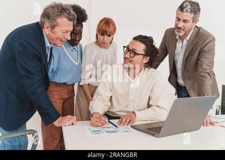Multicultural team, including senior and junior African and Caucasian members, gathers around young Chinese man's desk. He's explaining printouts of a Stock Photo