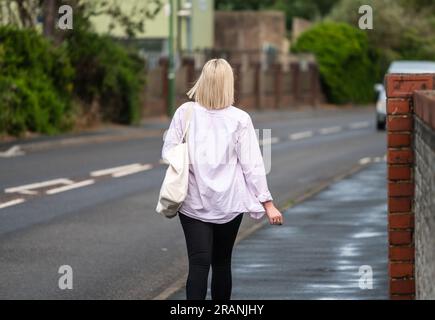 Rear of young classy lady with blonde hair, pink top and handbag walking away along a pavement in the UK. Stock Photo