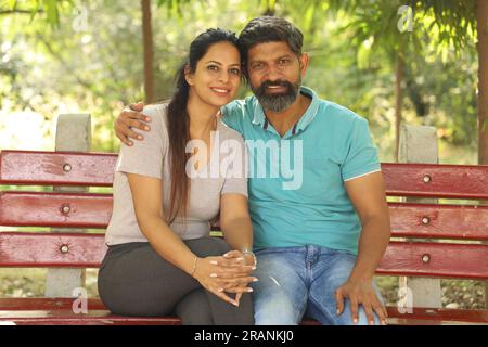 Portrait of a happy Indian couple sitting on the bench in the park amongst the greens. Stock Photo