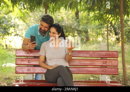 Portrait of a happy Indian couple sitting on the bench in the park amongst the greens. Having a video call. Couple looking into the smartphone Stock Photo