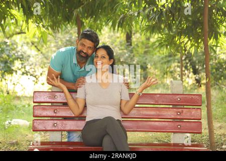 Portrait of a happy Indian couple sitting on the bench in the park amongst the greens. Stock Photo