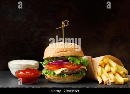 Juicy appetizing beef burger, french fries, two sauces on a black slate plate on a dark background. American fast food, homemade hamburger. Stock Photo