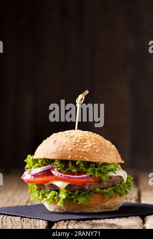 Appetizing burger with juicy beef cutlet, cheese, tomato, lettuce, onion on a wooden table and dark background. Fast food, homemade hamburger. Stock Photo
