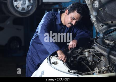 Mechanic in moustache repairing and examining the car. Car specialist is using repairing tools. Repairman wearing a blue mechanic's uniform. Stock Photo