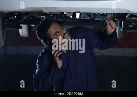 Mechanic working under lifted car in workshop. Specialist is examining under lifted car. Professional repairman is working hard in service station. Stock Photo