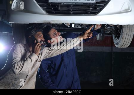 Car mechanics standing and working together in a service station. Car specialists examining the lifted car. Professional repairmen talking on phone. Stock Photo