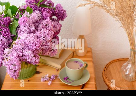 Vase with beautiful lilac flowers, books, cup of coffee on table and pampas grass in room, closeup Stock Photo