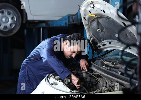 Close up portrait of hard working car mechanic standing in service station beside a car. Car Specialist is examining the engine. Stock Photo
