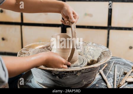 Cropped view of young female ceramicist making clay jug and working with pottery wheel near wooden tools in blurred art workshop , artisanal pottery p Stock Photo