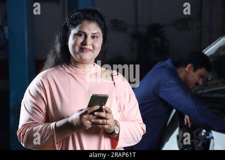 Happy customer at car workshop making online payment using smart phone, Hard working car mechanic standing behind the cusomer Stock Photo