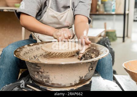 Cropped view of young female artisan in apron pouring water from sponge on clay on spinning pottery wheel near bowl in blurred art studio, pottery cre Stock Photo