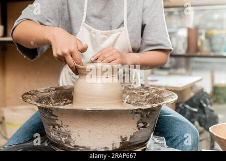 Cropped view of young female ceramicist in apron and workwear making shape of clay vase and working with pottery wheel in blurred ceramic workshop, po Stock Photo