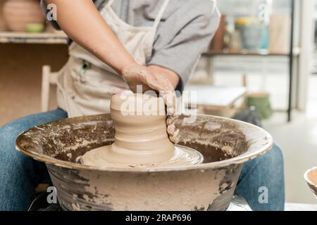 Cropped view of blurred female ceramicist in apron molding and shaping wet clay while working with spinning pottery wheel in art workshop, skilled pot Stock Photo