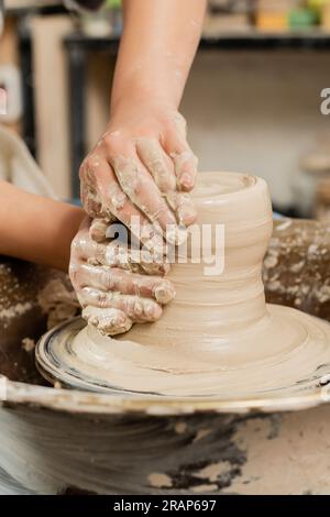 Cropped view of young female ceramicist molding wet clay on spinning pottery wheel while working in blurred ceramic workshop at background, skilled po Stock Photo