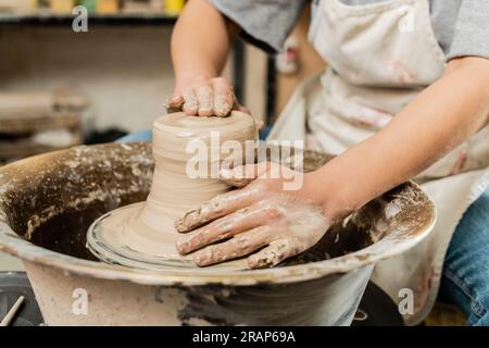 Cropped view of blurred female artisan in apron shaping wet clay and working with pottery wheel in ceramic art workshop at background, skilled pottery Stock Photo