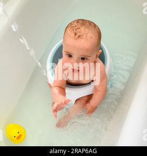 Baby boy in a baby bath in a full size bath fascinated by running water with yellow plastic duck Stock Photo