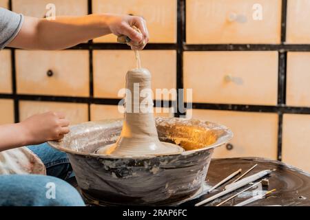 Cropped view of young female potter pouring water from sponge on clay while working on pottery wheel near tools in workshop at background, artisan cre Stock Photo
