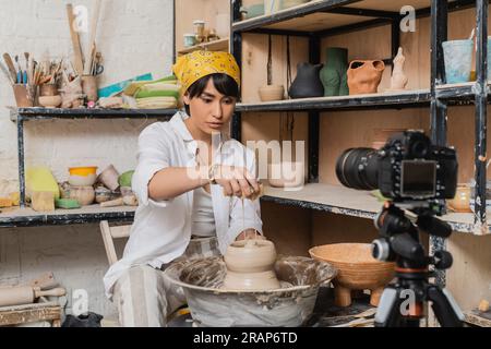 Young asian female potter in headscarf pouring water from sponge on clay on pottery wheel near blurred digital camera in ceramic workshop, clay sculpt Stock Photo