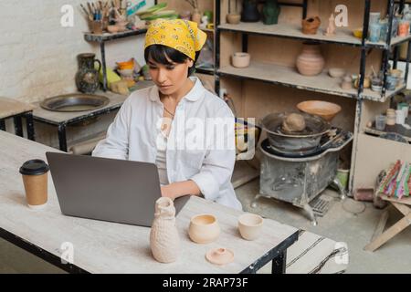 Young brunette asian craftswoman in headscarf and workwear using laptop while working near coffee to go and clay products on table in ceramic workshop Stock Photo